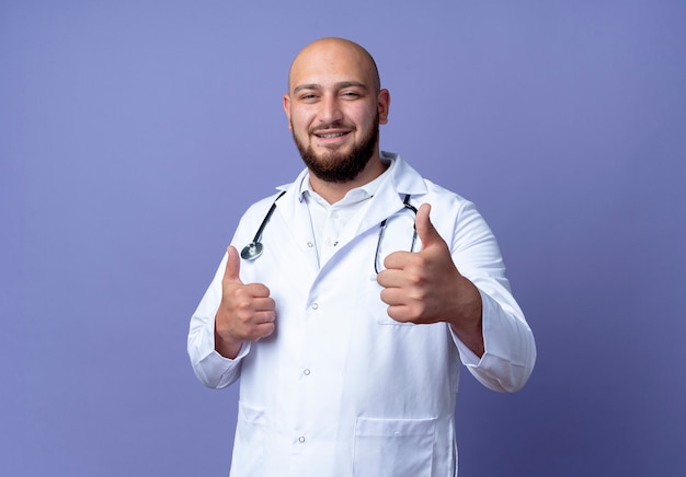 Foto gratuita satisfecho joven médico varón calvo vistiendo bata médica y estetoscopio con el pulgar hacia arriba aislado sobre fondo azul.