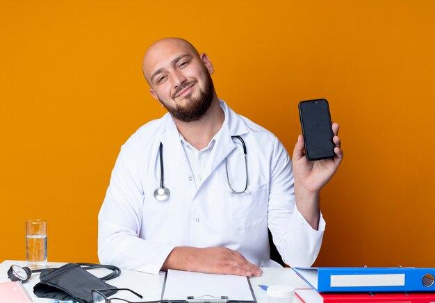 Satisfecho joven médico varón calvo vistiendo bata médica y estetoscopio sentado en la mesa de trabajo con herramientas médicas sosteniendo teléfono aislado sobre fondo naranja