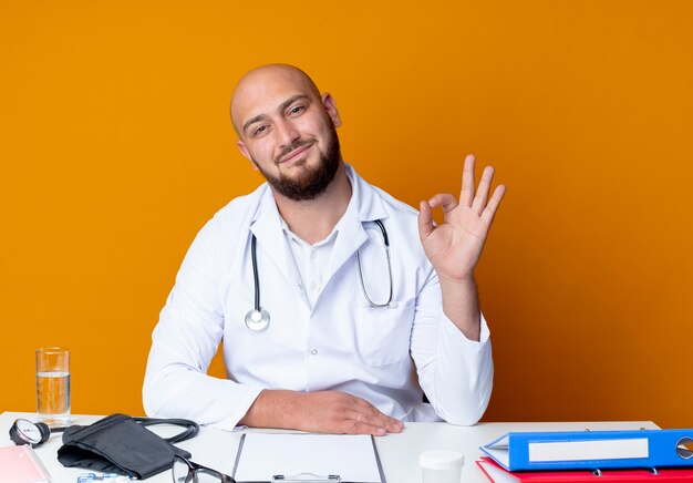 Satisfecho joven médico varón calvo vistiendo bata médica y estetoscopio sentado en la mesa de trabajo