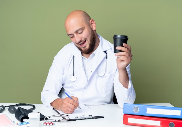 Foto gratuita satisfecho joven médico varón calvo vistiendo bata médica y un estetoscopio sentados frente al escritorio