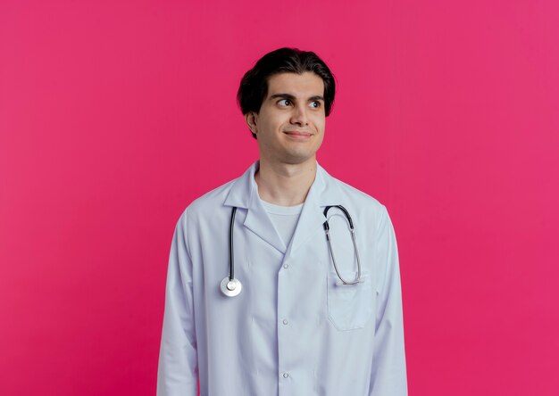 Satisfecho joven médico vistiendo bata médica y un estetoscopio mirando al lado aislado en la pared rosa con espacio de copia