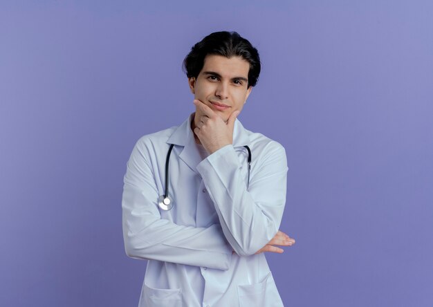 Satisfecho joven médico vistiendo bata médica y estetoscopio poniendo la mano en la barbilla aislada en la pared púrpura con espacio de copia