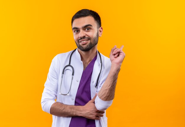 Satisfecho joven médico vistiendo estetoscopio bata médica apunta hacia atrás con el dedo sobre fondo amarillo aislado