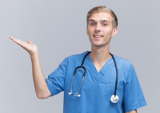 Satisfecho joven médico vistiendo uniforme médico con puntos de estetoscopio con la mano al lado aislado en la pared blanca con espacio de copia