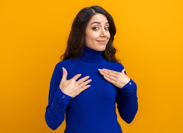 Satisfecho joven mujer bonita apuntando a sí misma mirando al frente aislado en la pared naranja