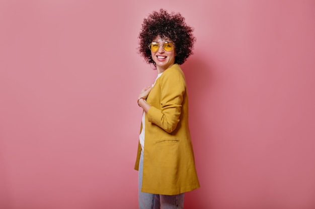 Satisfecho joven sonriente con rizos vistiendo chaqueta amarilla mirando al frente sobre pared rosa