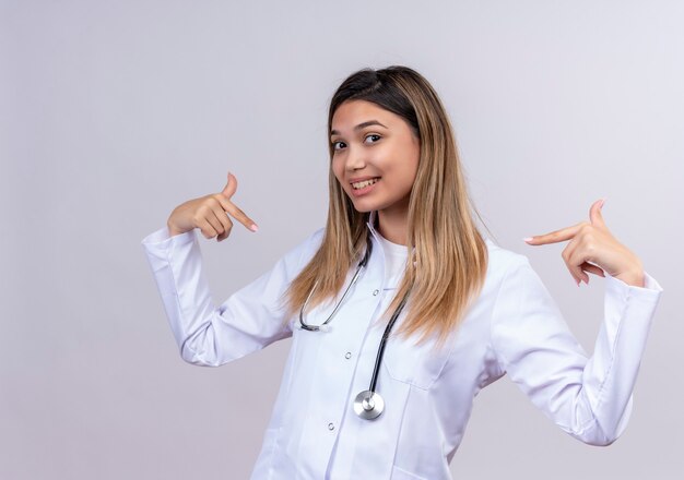 Satisfecho médico joven y bella mujer vistiendo bata blanca con estetoscopio apuntando con el dedo índice a sí misma mirando confiada