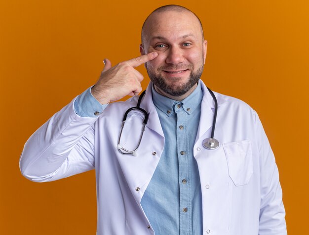 Satisfecho médico varón de mediana edad vistiendo bata médica y estetoscopio mirando al frente poniendo el dedo debajo del ojo aislado en la pared naranja