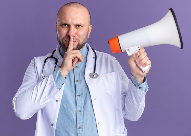 Satisfecho médico varón de mediana edad vistiendo bata médica y estetoscopio sosteniendo altavoz haciendo gesto de silencio aislado en la pared púrpura