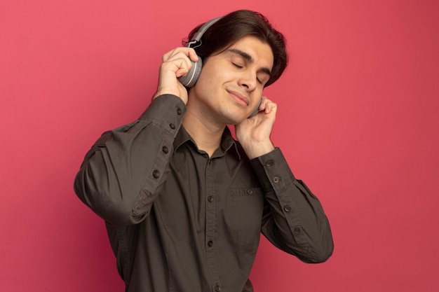 Satisfecho con los ojos cerrados, chico guapo joven con camiseta negra con auriculares escucha música aislada en la pared rosa