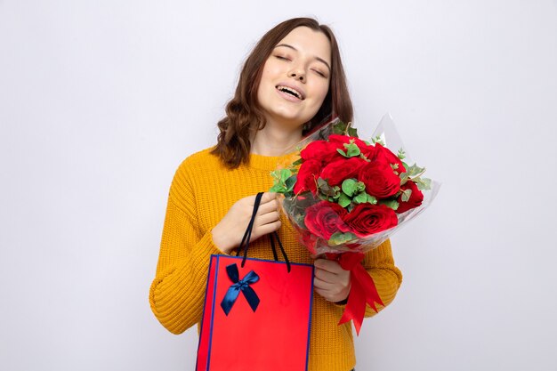 Satisfecho con los ojos cerrados hermosa joven en el día de la mujer feliz con bolsa de regalo con ramo aislado en la pared blanca