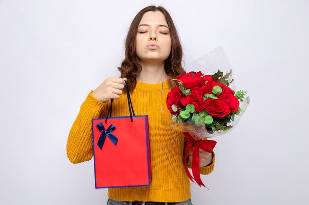 Satisfecho con los ojos cerrados hermosa joven en el día de la mujer feliz con bolsa de regalo con ramo aislado en la pared blanca