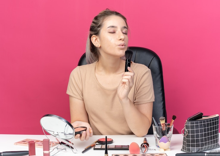 Foto gratuito satisfecho con los ojos cerrados, la hermosa joven se sienta a la mesa con herramientas de maquillaje aplicando rubor en polvo aislado en la pared rosa