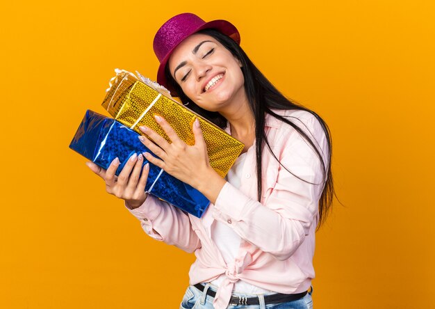 Satisfecho con los ojos cerrados hermosa joven vistiendo gorro de fiesta sosteniendo cajas de regalo