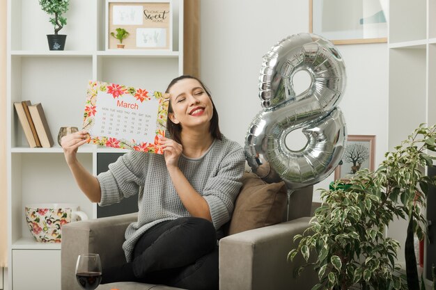 Satisfecho con los ojos cerrados hermosa mujer en el día de la mujer feliz celebración de calendario sentado en un sillón en la sala de estar