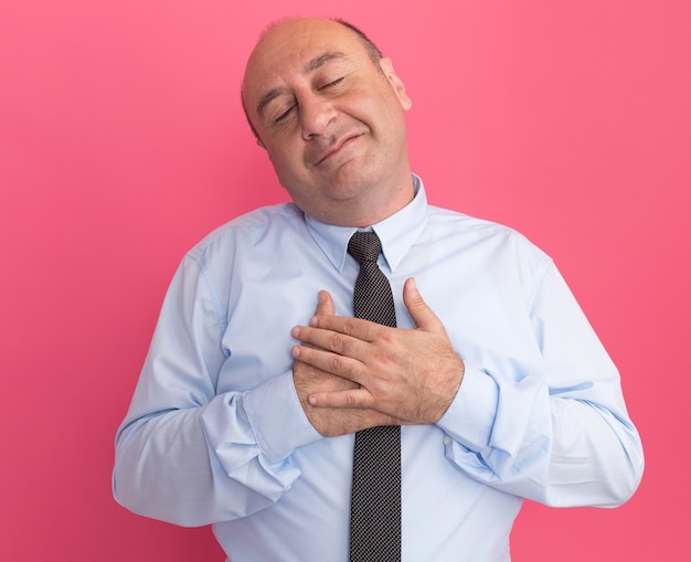 Satisfecho con los ojos cerrados hombre de mediana edad vestido con camiseta blanca con corbata poniendo la mano en el corazón aislado en la pared rosa