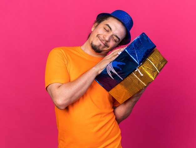 Satisfecho con los ojos cerrados inclinando la cabeza joven con sombrero de fiesta sosteniendo cajas de regalo aisladas en la pared rosa