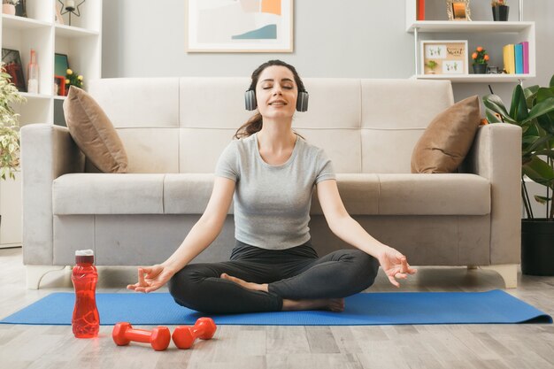 Satisfecho con los ojos cerrados joven con audífonos haciendo ejercicio en la estera de yoga frente al sofá en la sala de estar