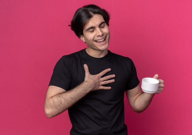 Foto gratuita satisfecho con los ojos cerrados joven guapo con camiseta negra sosteniendo una taza de café aislado en la pared rosa