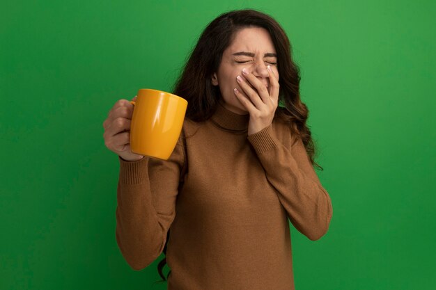 Satisfecho con los ojos cerrados joven hermosa sosteniendo una taza de té y la boca cubierta con la mano aislada en la pared verde