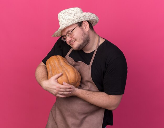 Satisfecho con los ojos cerrados joven jardinero con sombrero de jardinería abrazó calabaza aislado en la pared rosa