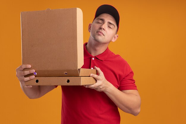 Satisfecho con los ojos cerrados, joven repartidor vestido con uniforme con apertura de tapa y oliendo caja de pizza aislada en la pared naranja