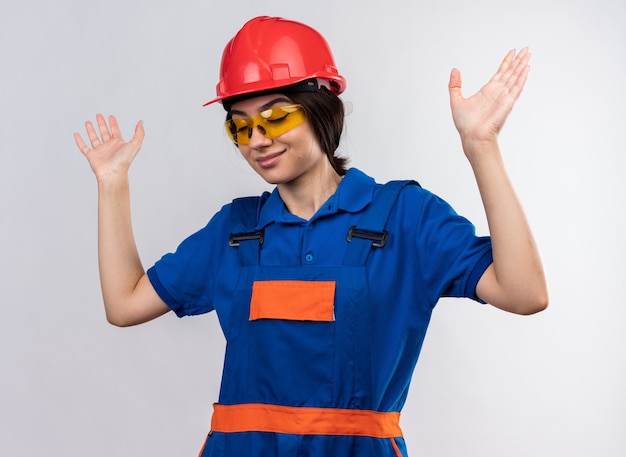 Satisfecho con los ojos cerrados mujer joven constructora en uniforme y gafas levantando las manos aisladas en la pared blanca