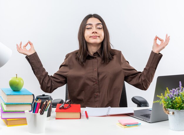 Satisfecho con los ojos cerrados, la mujer joven de la escuela se sienta a la mesa con herramientas escolares haciendo gesto de meditación