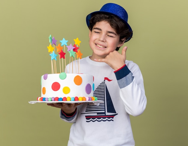 Satisfecho con los ojos cerrados niño vistiendo gorro de fiesta azul sosteniendo pastel mostrando gesto de llamada telefónica