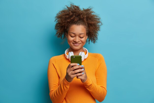 Satisfecho suscriptor afroamericano adicto a las redes sociales y las tecnologías modernas sostiene mensajes de texto de tipo celular usa auriculares estéreo alrededor del cuello vestida con ropa casual
