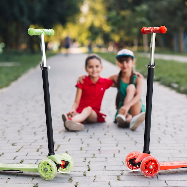 Foto gratuita scooters de patada roja y verde frente a dos chicas sentadas juntas en la pasarela