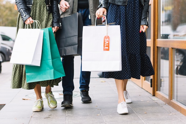 Sección baja de amigos caminando por la acera con coloridas bolsas de compras.