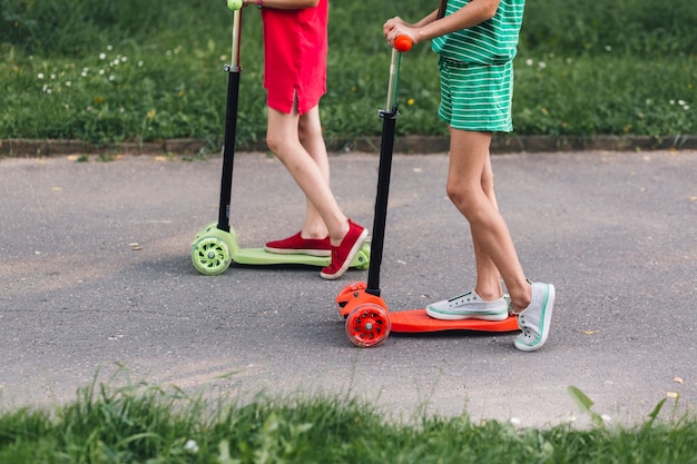 Sección baja de dos chicas montando patinete en el parque