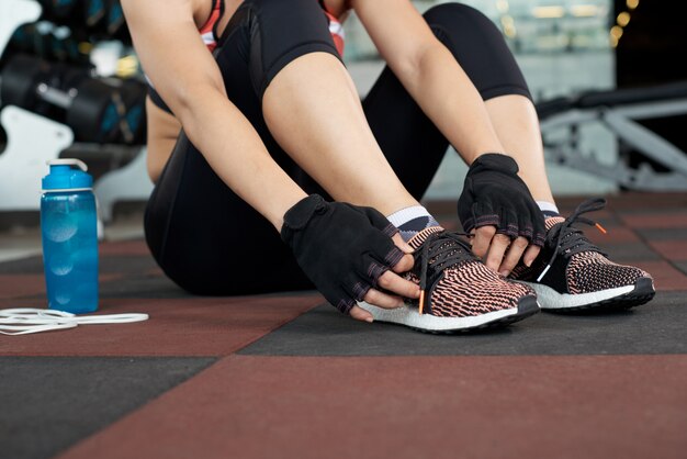 Sección baja de mujer irreconocible en ropa deportiva sentada en el piso del gimnasio atando cordones y preparándose para el entrenamiento