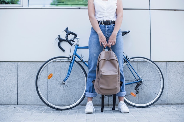 Foto gratuita sección baja de la mujer joven que sostiene la mochila en su mano que se opone a la bicicleta