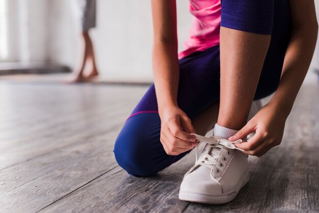 Sección baja de una niña atándose cordones de zapatos blancos.