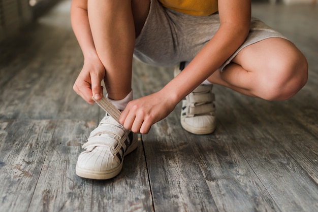 Sección baja de un niño que pone la correa del zapato en el piso de madera dura