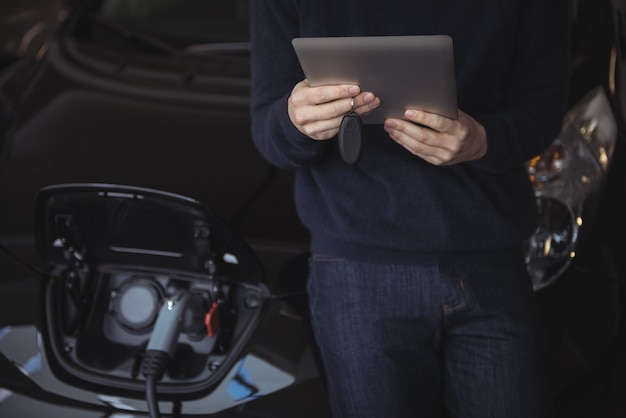 Sección intermedia del hombre con tableta digital mientras se carga un coche eléctrico