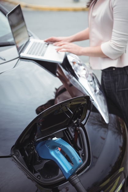 Sección intermedia de mujer con portátil mientras carga un coche eléctrico