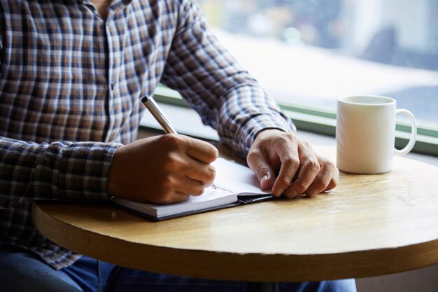 Sección media del hombre anónimo en camisa a cuadros escribiendo notas en la mesa de café