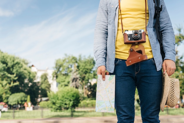 Foto gratuita sección media del hombre turista que lleva la cámara y que sostiene el mapa y el sombrero en el parque