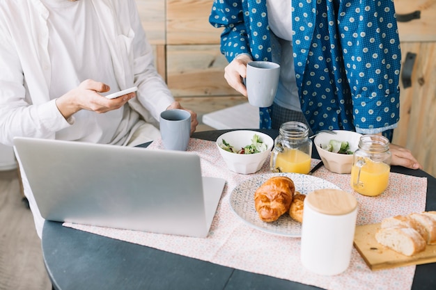 Sección media de hombres sosteniendo una taza de café cerca de un delicioso desayuno con jugo y una computadora portátil sobre una mesa de madera