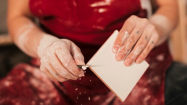 Sección media de la mano del alfarero femenino que quita la pintura en el borde del azulejo