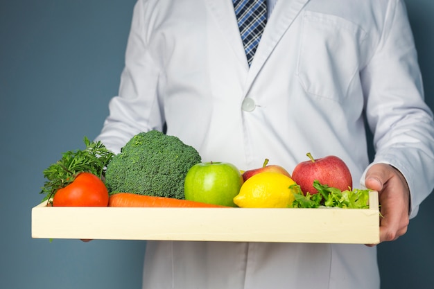 Sección media de un médico con bandeja de madera llena de verduras y frutas saludables sobre fondo gris