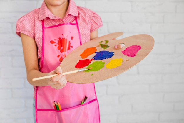 Sección media de una niña con delantal rosa que mezcla la pintura en la paleta con pincel