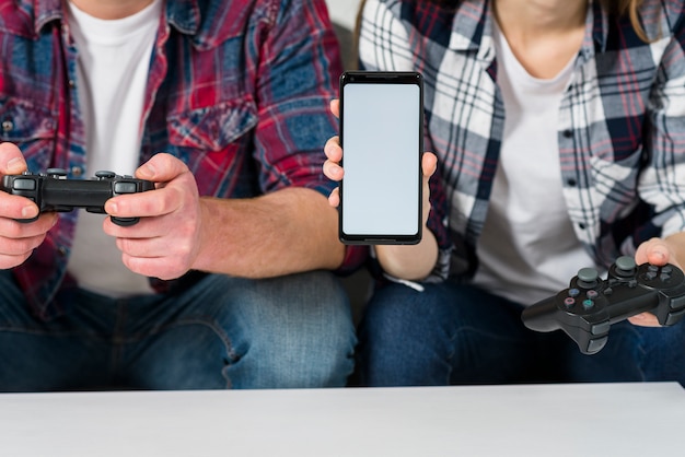 Sección media de una pareja joven jugando videojuegos que muestra un teléfono inteligente con pantalla en blanco