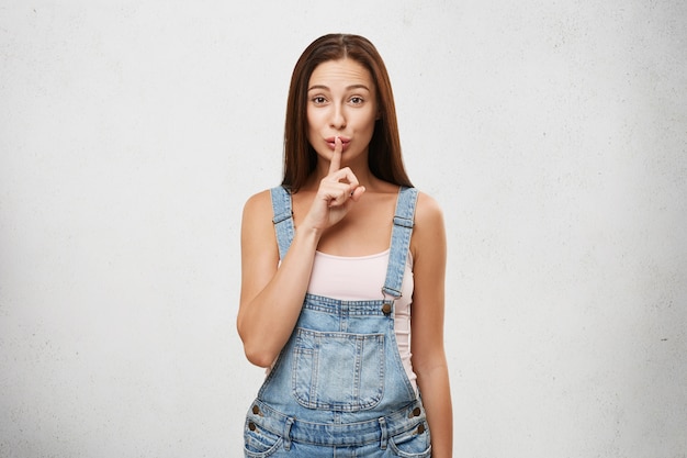 Secreto, chismes de conspiración e información segura. Imagen de una hermosa mujer morena con elegante mono de jeans, sosteniendo el dedo en los labios como un gesto silencioso, pidiendo guardar silencio sobre su secreto