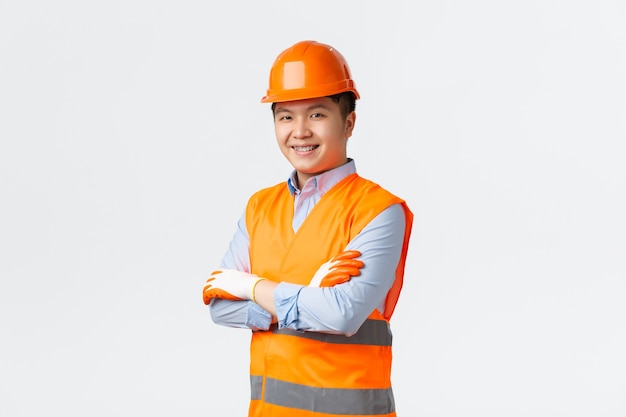 Sector de la construcción y concepto de trabajadores industriales. Confiado joven ingeniero asiático, gerente de construcción con ropa reflectante y casco, brazos cruzados y una sonrisa descarada, asegurando calidad, pared blanca