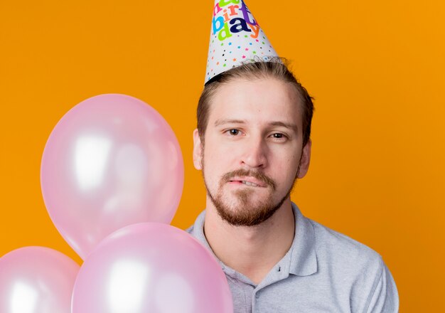 Seductor joven en gorra de fiesta sosteniendo un montón de globos de aire mordiendo el labio concepto de fiesta de cumpleaños de pie sobre la pared naranja