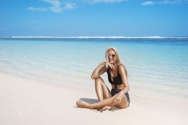 Seductora feliz sonriente rubia bronceada turista disfrutar de vacaciones Maldivas islas paradisíacas sentarse con las piernas cruzadas playa de arena usar pantalones cortos gafas de sol relajarse cerca de la costa vista al mar viajar verano